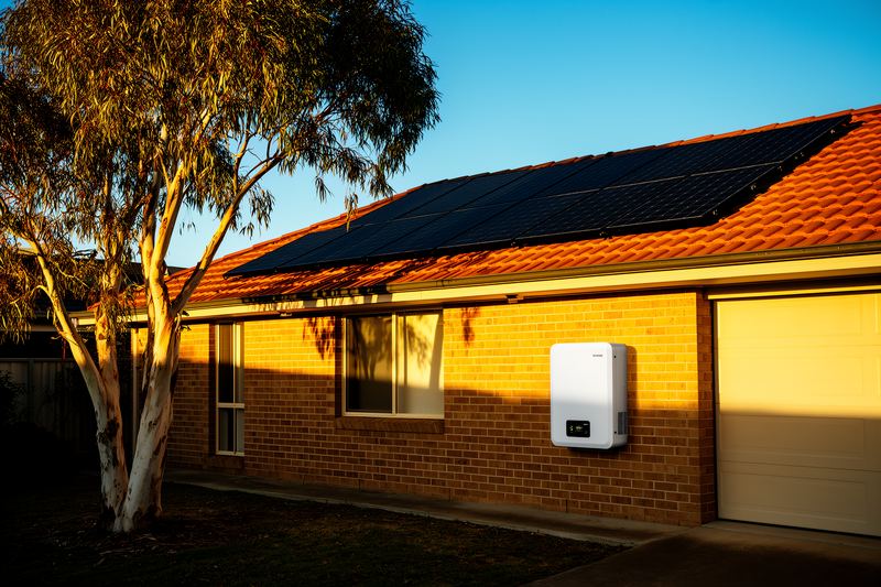 Brick house with black solar panels on a terracotta roof and a white home battery mounted on the wall beside the garage